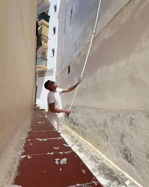 Pintor pintando con pértiga la pared alta de un callejón entre dos edificios